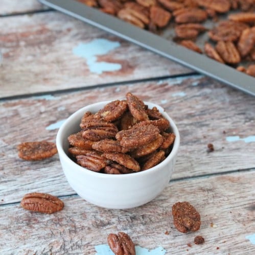 Cajun pecans in a white bowl with pecans on a tray behind it