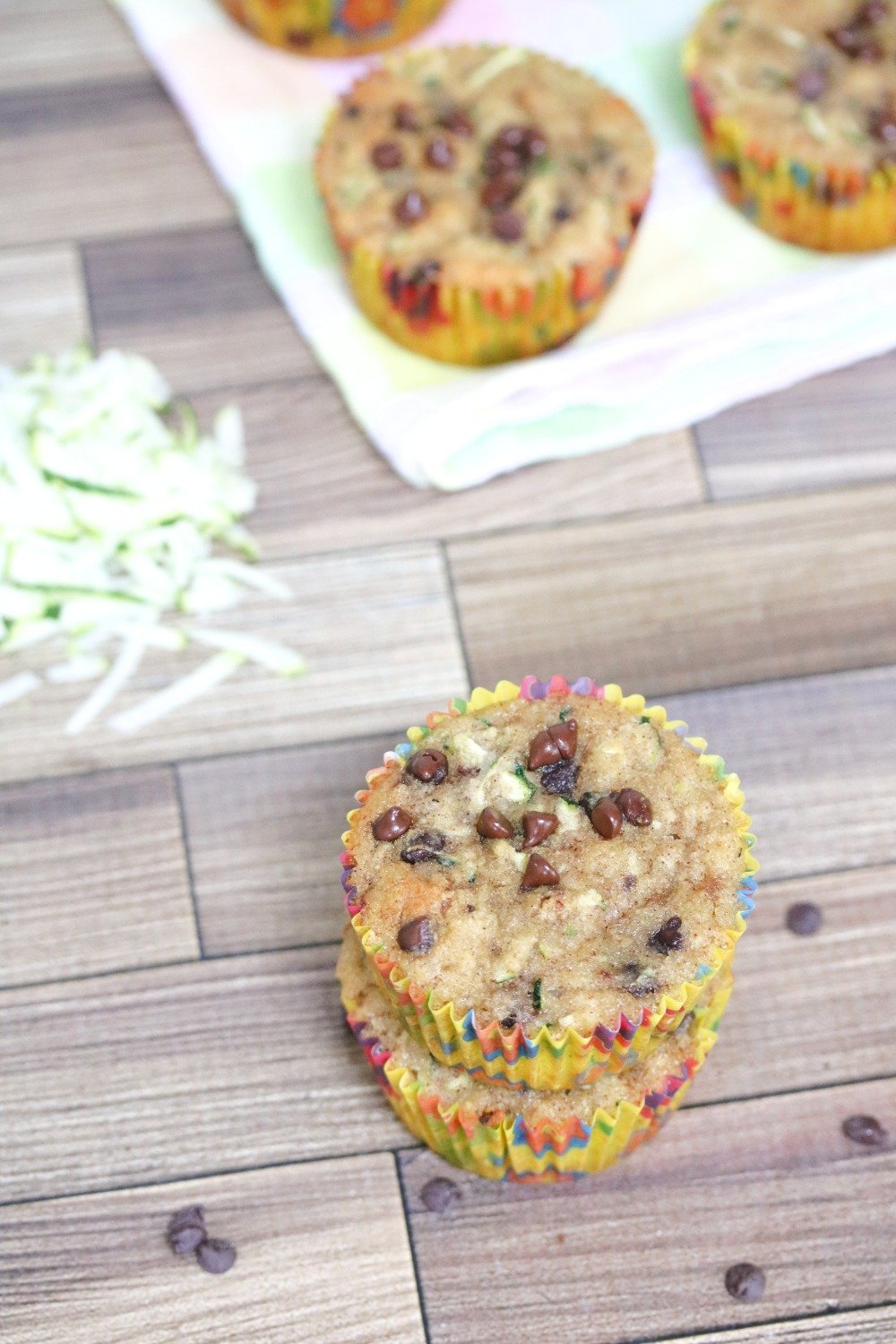 Two chocolate chip muffins stacked with shredded zucchini behind them and more muffins on a linen
