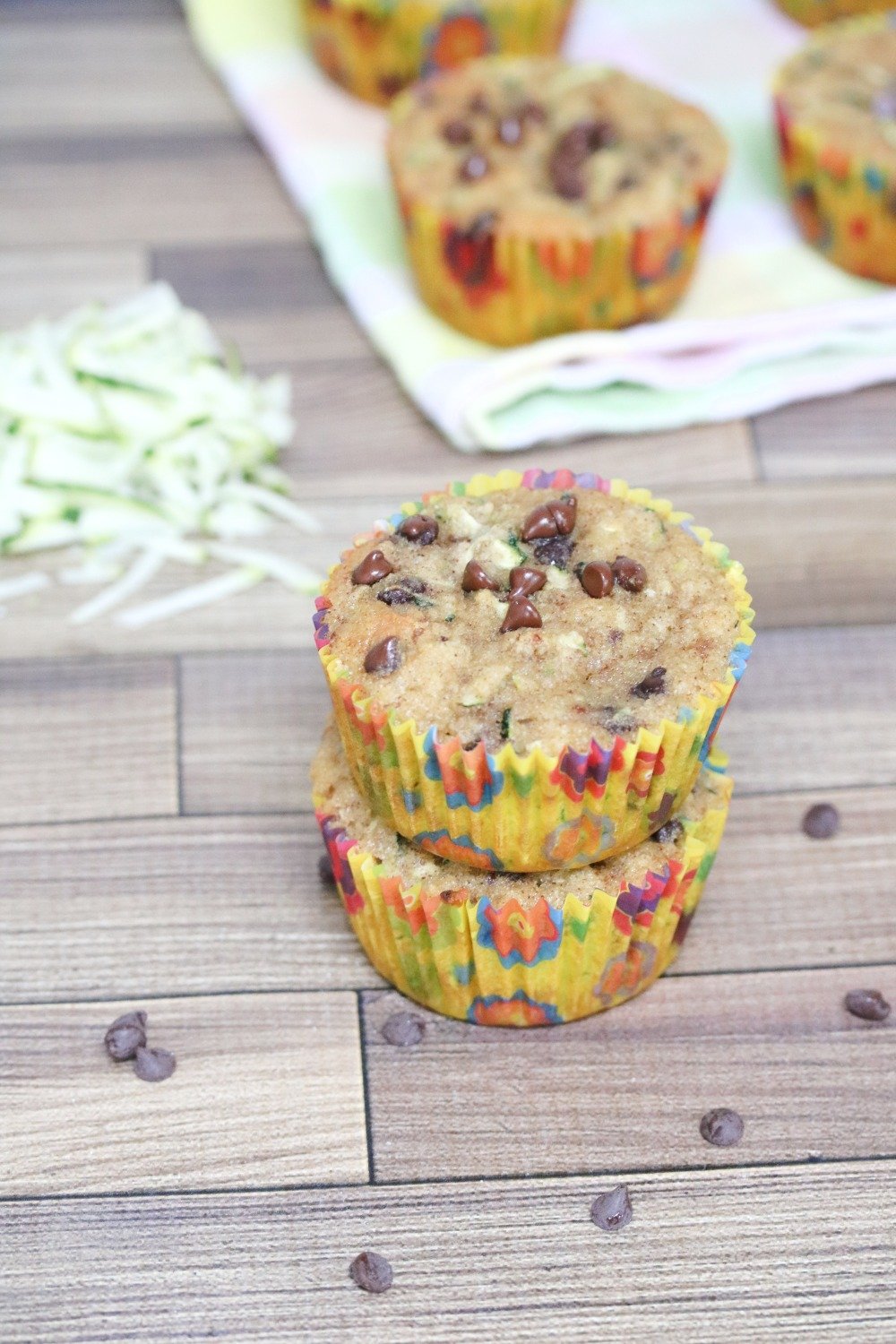 Two chocolate chip muffins stacked with shredded zucchini behind them and more muffins on a linen