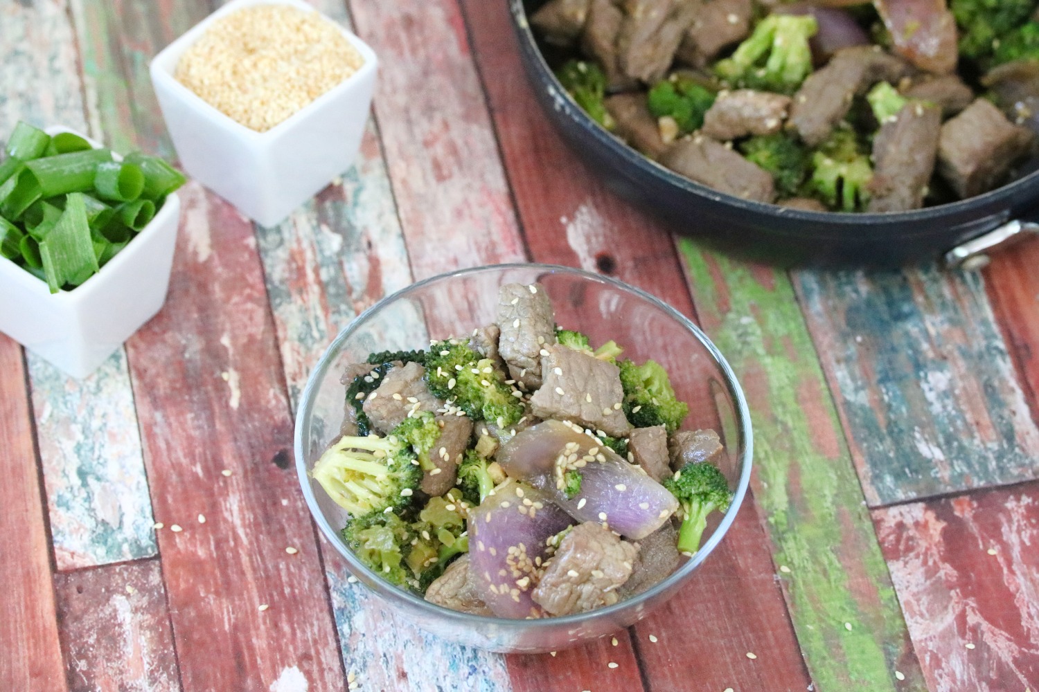 Beef and broccoli stir fry topped with sesame seeds in a glass bowl with sesame seeds and green onions in white dishes beside it and a pan with the remaining stir fry behind it