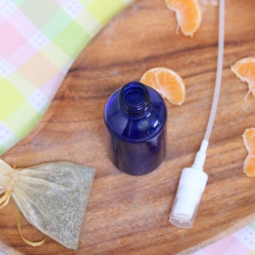 Blue glass bottle on a wooden cutting board on top of a colored linen with spray nozzle beside it. Orange slices and a bag of lavender around beside the spray bottle.
