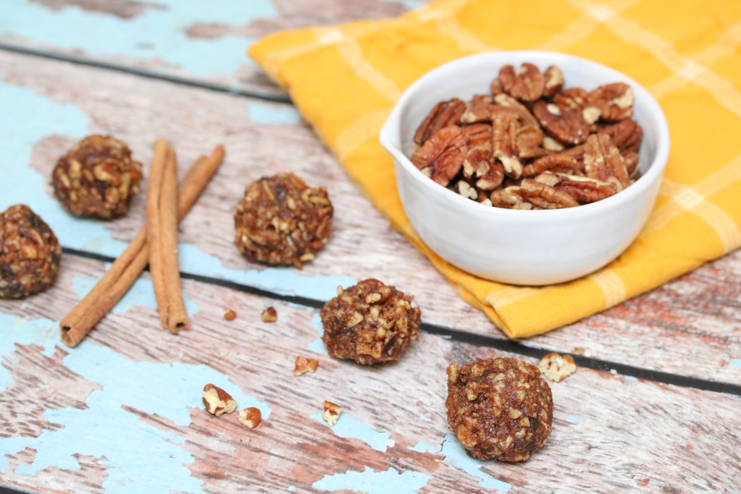 Three no-bake pecan balls in front of a bowl with pecans on a yellow cloth and cinnamon sticks behind it