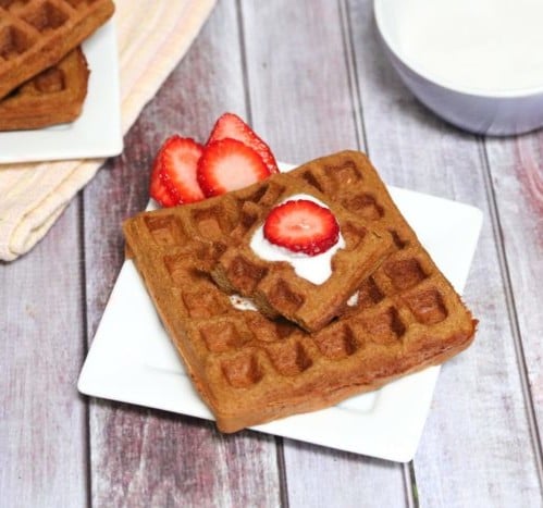 Square gingerbread waffle on a white plate with coconut milk and strawberries on top and more waffles and coconut milk behind it