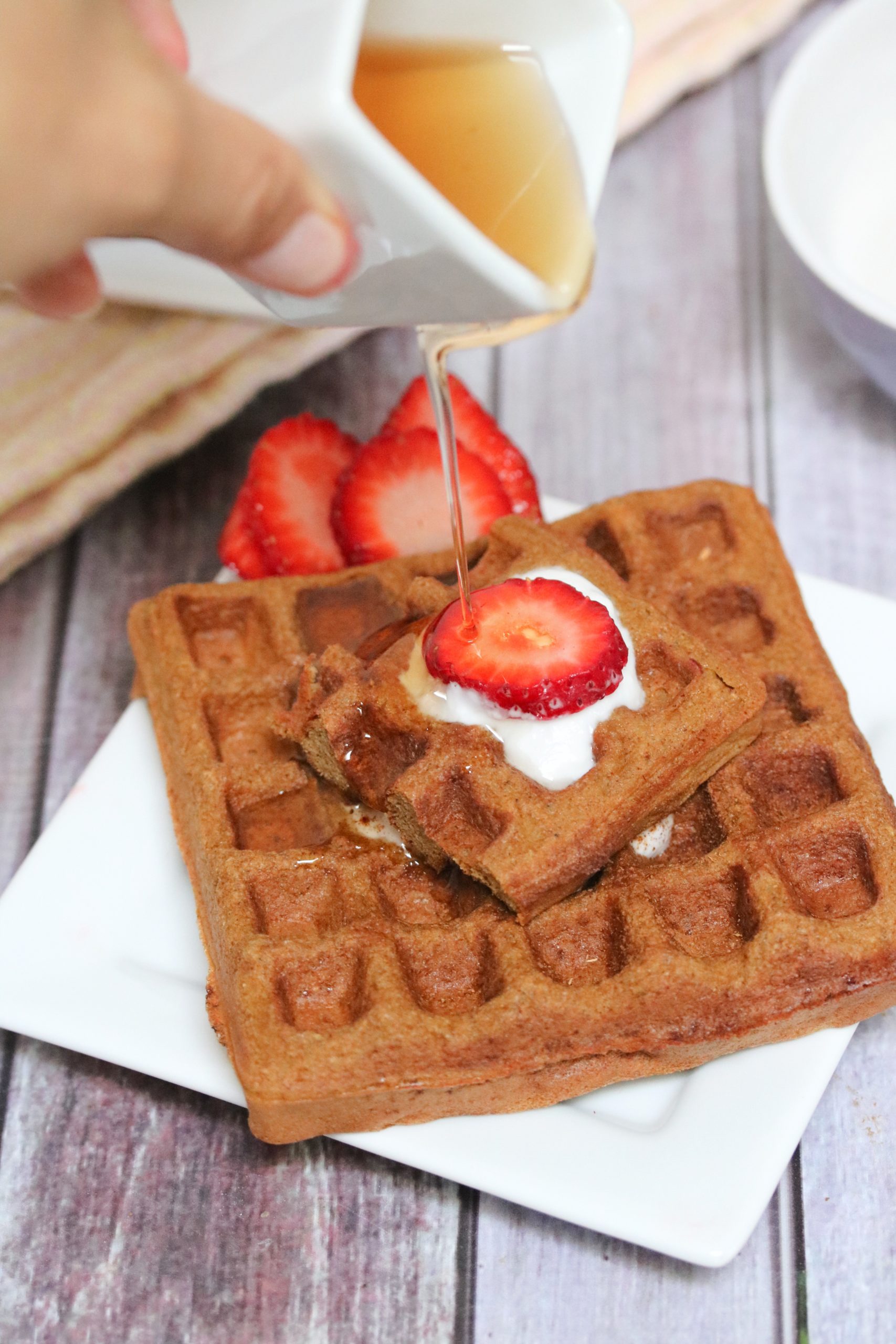 Square gingerbread waffle on a white plate with coconut milk and strawberries on top and more waffles and coconut milk behind it